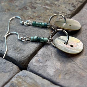 May include: A pair of dangle earrings featuring silver-toned metal hooks connected to a short strand of small, cylindrical teal beads. Each earring terminates in a circular, iridescent, off-white button with two central holes suspended by a silver ring.