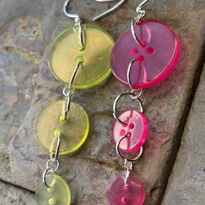 May include: Dangling earrings featuring a series of translucent buttons in shades of yellow-green and pink. The buttons are connected by silver-colored metal rings and hooks, creating a layered, eye-catching design. The earrings are displayed against a textured, gray stone surface.