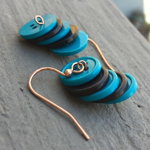 May include: Teal and black button earrings with a rose gold hook. The buttons are stacked on top of each other.
