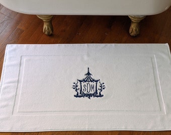 May include: White bath mat with a navy blue monogram in the center. The monogram is a stylized design with a tent-like structure and the letters "SDM" inside.