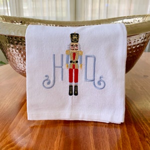 May include: White kitchen towel with a grey embroidered monogram "JHD" and a red and gold embroidered nutcracker in the centre.