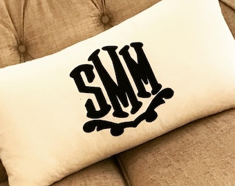 Op de afbeelding: Crèmekleurig rechthoekig kussen met de zwarte letters "SMM" in een decoratief lettertype. Het kussen ligt op een beige gecapitonneerde bank. Een decoratief woonaccessoire.