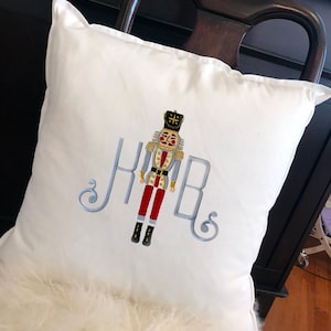 May include: White pillow with embroidered nutcracker design and the monogram "KHB" in blue thread.