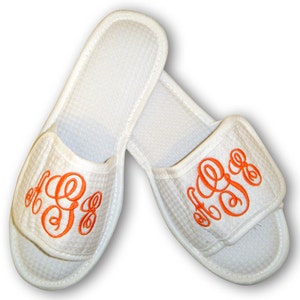 Puede incluir: Un par de zapatillas blancas de tejido tipo gofre con monogramas bordados en naranja. Las zapatillas tienen la punta cerrada y el talón abierto.