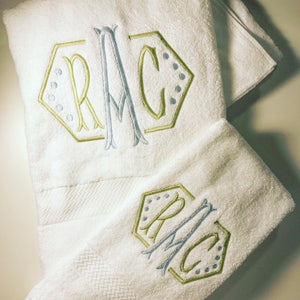 May include: Two white towels with embroidered monograms in a geometric shape. The monogram is in blue and green thread. The towels are folded and stacked on top of each other.