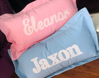 Op de afbeelding: Twee gepersonaliseerde kussenslopen, een roze met de naam "Eleanor" in witte cursieve letters en een lichtblauwe met de naam "Jaxon" in witte blokletters.