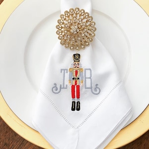 May include: White linen napkin with a gold jeweled napkin ring and an embroidered nutcracker design with the monogram "JRB".