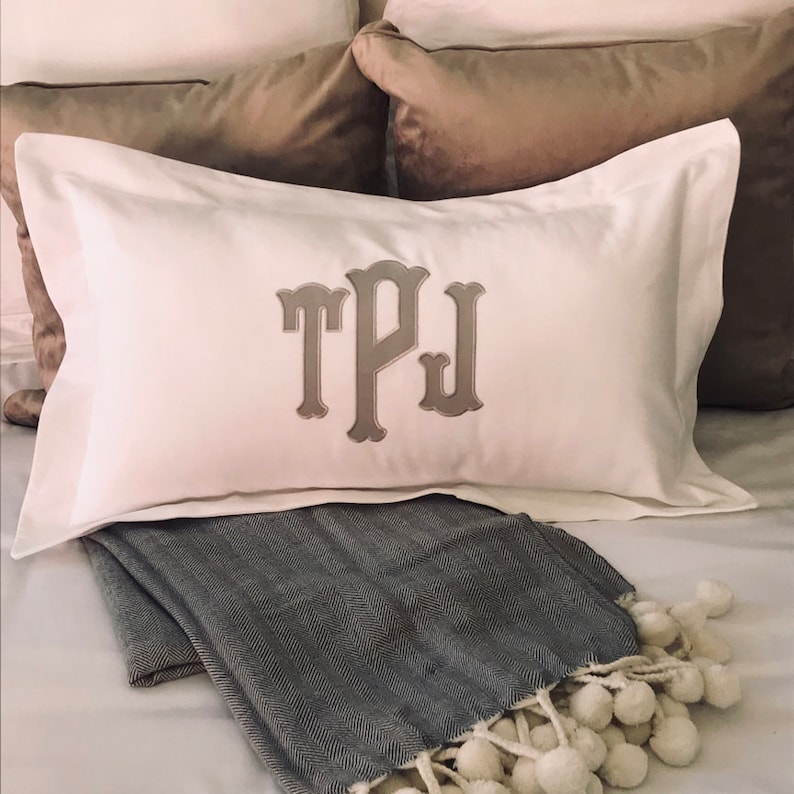 May include: A white pillow with a gray embroidered monogram "TPJ" on a bed with a gray and white throw blanket.