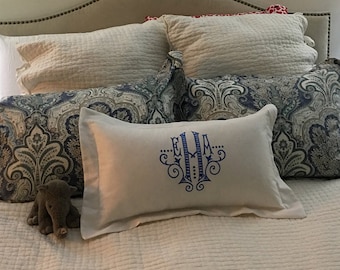 Op de afbeelding: Een wit bed met een wit hoofdeinde en een wit dekbed. Er liggen verschillende kussens op het bed, waaronder een wit kussen met blauwe borduurwerk dat "E.M.A." zegt.
