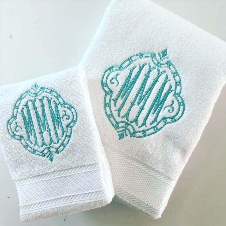 May include: Two white towels with a teal monogram of the letters MMM in a decorative frame. The towels are folded and displayed on a white surface.