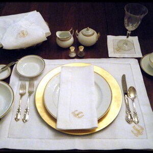 May include: A white monogrammed table setting with a gold rimmed plate, white napkin, and silver cutlery. There are two small white creamer and sugar containers with gold accents.