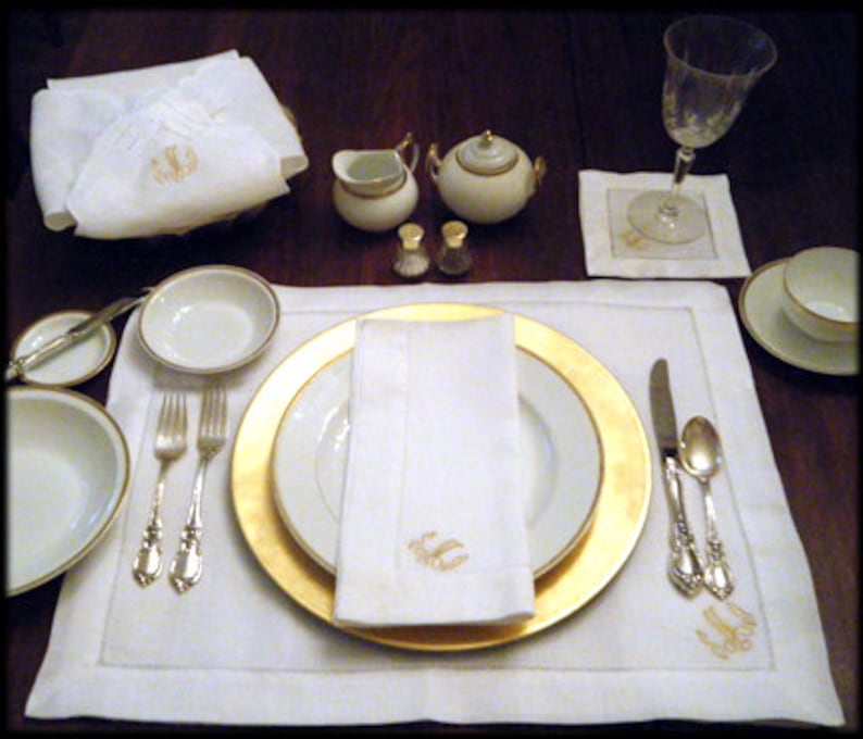 May include: A white monogrammed table setting with a gold rimmed plate, white napkin, and silver cutlery. There are two small white creamer and sugar containers with gold accents.