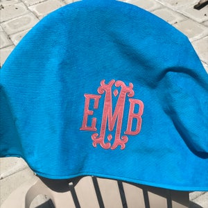 May include: A bright turquoise towel with a pink embroidered monogram of the letters E, M, and B. The towel is folded over a chair, showcasing the soft texture and vibrant color. The monogram is in a decorative, flowing font.