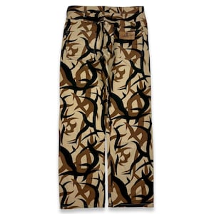 May include: A pair of tan and black camouflage pants with a tribal pattern. The pants have a brown leather patch on the back with the text "Fickle Apparel" in white.