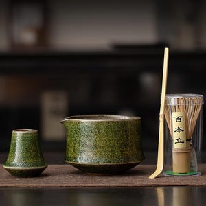 May include: A matcha tea set comprising a green ceramic bowl, a small cup, a bamboo whisk, and a bamboo scoop. The whisk is in a clear container with Japanese text. The set is displayed on a brown surface.
