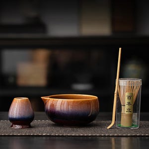 May include: A ceramic matcha bowl and a matching whisk holder featuring a gradient glaze transitioning from deep purple at the base to a warm amber at the rim. A bamboo whisk is stored in a clear plastic container with the Japanese text 百本立.