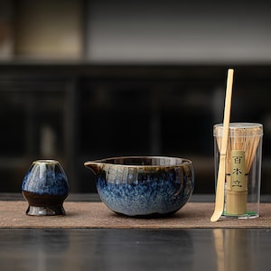 May include: A ceramic tea set with a blue and brown glaze. The set includes a small tea bowl, a larger bowl with a spout, a bamboo whisk, and a bamboo scoop. The whisk is in a clear container with Japanese text.