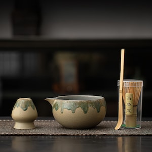Puede incluir: Un juego de té matcha con un cuenco de cerámica con pico, una pequeña taza de cerámica, un batidor de bambú y una cuchara de bambú. Las piezas de cerámica tienen una base beige con esmalte verde goteando. El batidor está en un recipiente de plástico transparente.