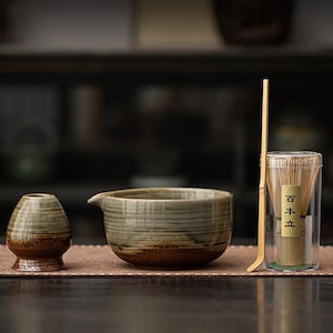 May include: A matcha tea set comprising a ceramic bowl, a whisk, a small ceramic container, and a bamboo scoop. The bowl and container have a brown and green colour scheme. The whisk is in a clear container with Japanese text.