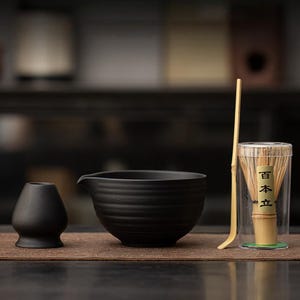 May include: A black ceramic matcha bowl with a spout, a whisk holder, and a bamboo whisk in a clear case. A bamboo scoop is also included. The items are arranged on a brown mat, ideal for preparing matcha green tea.