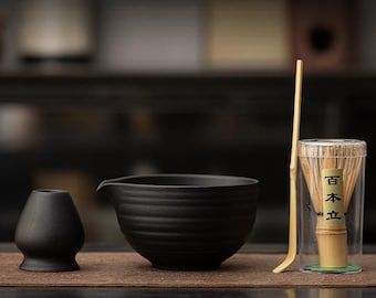 Può includere: Un set da matcha nero con una ciotola, un supporto, un frullino di bambù in una custodia trasparente e un cucchiaio di bambù. Gli articoli sono disposti su un tappetino marrone.
