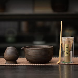 Puede incluir: Un juego de té matcha con un cuenco de cerámica marrón con boquilla, un pequeño soporte para batidor de cerámica, un batidor de bambú y un recipiente con un batidor de bambú. El juego está sobre una esterilla marrón.