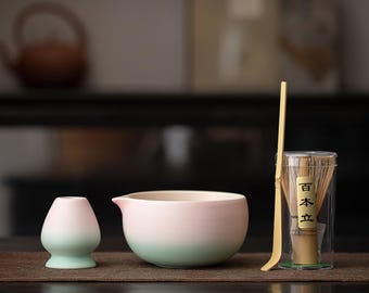 Puede incluir: Un juego de té matcha con un cuenco de cerámica degradado rosa y verde, un chawan a juego, un batidor de bambú y una cuchara. El batidor está en un recipiente transparente con texto japonés. El juego está dispuesto sobre una esterilla marrón.