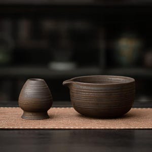 K&ouml;nnte beinhalten: Ein braunes Keramik-Teeschalen- und Quirl-Set. Die Schale hat eine breite, runde Form mit einem Ausguss und horizontalen Rillen. Der Quirl ist ein kleines, vasenf&ouml;rmiges Objekt mit rundem Boden und horizontalen Rillen. Das Set steht auf einer braunen Matte.