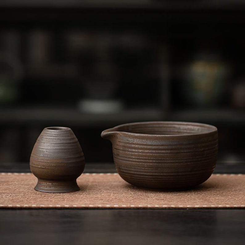 K&ouml;nnte beinhalten: Ein braunes Keramik-Teeschalen- und Quirl-Set. Die Schale hat eine breite, runde Form mit einem Ausguss und horizontalen Rillen. Der Quirl ist ein kleines, vasenf&ouml;rmiges Objekt mit rundem Boden und horizontalen Rillen. Das Set steht auf einer braunen Matte.