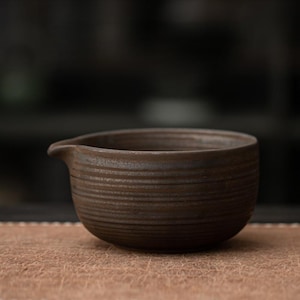 K&ouml;nnte beinhalten: Eine kleine, braune Keramikschale mit Ausguss. Die Schale hat ein strukturiertes, horizontal gestreiftes Design. Die dunkelbraune Farbe und das schlichte Design deuten auf eine rustikale oder minimalistische &Auml;sthetik hin. Die Schale wird wahrscheinlich zum Servieren oder Ausgie&szlig;en verwendet.