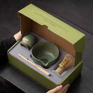 K&ouml;nnte beinhalten: Ein Matcha-Teeset in einer gr&uuml;nen Box. Das Set enth&auml;lt eine Keramikschale, einen Schneebesen, einen L&ouml;ffel und einen kleinen Beh&auml;lter. Schale und Beh&auml;lter haben eine gr&uuml;ne und braune Glasur. Die Box hat Text auf Englisch und Japanisch.