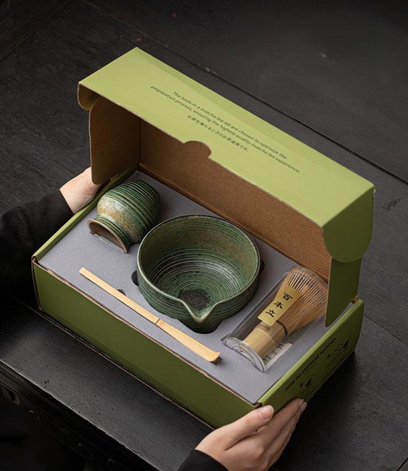 K&ouml;nnte beinhalten: Ein Matcha-Teeset in einer gr&uuml;nen Box. Das Set enth&auml;lt eine Keramikschale, einen Schneebesen, einen L&ouml;ffel und einen kleinen Beh&auml;lter. Schale und Beh&auml;lter haben eine gr&uuml;ne und braune Glasur. Die Box hat Text auf Englisch und Japanisch.
