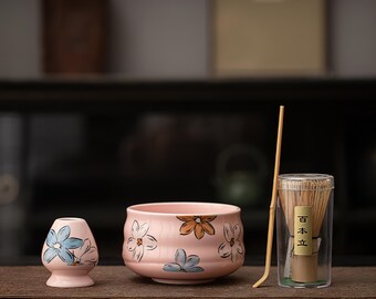 May include: A pink ceramic matcha tea set with floral designs. The set includes a bowl, whisk holder, bamboo whisk, and a bamboo scoop. The floral designs are in shades of blue, orange, and white.