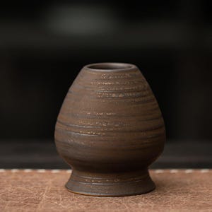 K&ouml;nnte beinhalten: Eine braune Matcha-Schale aus Keramik mit breitem Boden und schmaler &Ouml;ffnung. Die Schale hat eine strukturierte, geriffelte Oberfl&auml;che und eine dunkelbraune Farbe. Die Schale ist zum Aufschlagen von Matcha-Tee konzipiert und hat eine traditionelle Form.