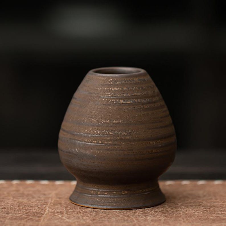 K&ouml;nnte beinhalten: Eine braune Matcha-Schale aus Keramik mit breitem Boden und schmaler &Ouml;ffnung. Die Schale hat eine strukturierte, geriffelte Oberfl&auml;che und eine dunkelbraune Farbe. Die Schale ist zum Aufschlagen von Matcha-Tee konzipiert und hat eine traditionelle Form.