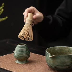 K&ouml;nnte beinhalten: Ein Matcha-Besen aus Bambus, oder Chasen, wird &uuml;ber einer kleinen, gr&uuml;nen Keramikschale und einer passenden Matcha-Schale gehalten. Der Schneebesen besteht aus d&uuml;nnen Bambusstreben, die zusammengebunden sind. Die Schalen haben ein strukturiertes, geripptes Design. Die Szene deutet auf eine Teezeremonie hin.