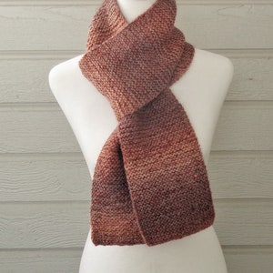 May include: A brown and pink knitted scarf with a gradient pattern is displayed on a white mannequin.
