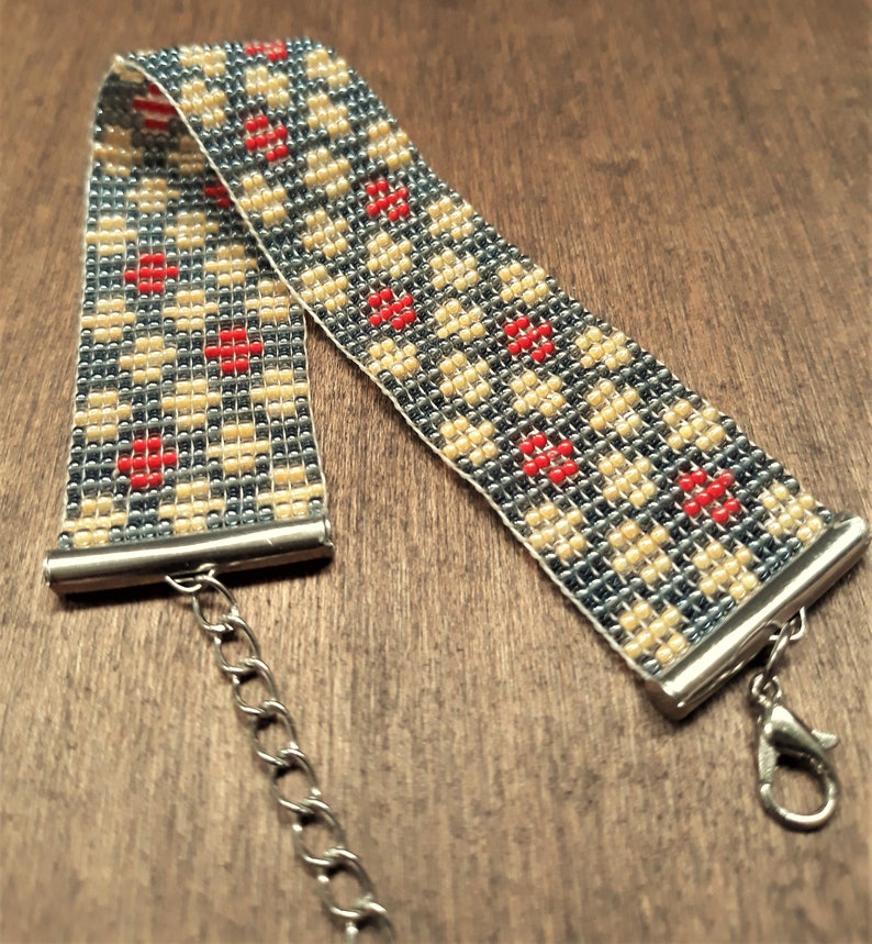 Peut inclure: Un bracelet en perles avec un motif g&eacute;om&eacute;trique dans les tons de gris, beige et rouge. Le bracelet a un fermoir et une cha&icirc;ne argent&eacute;s.