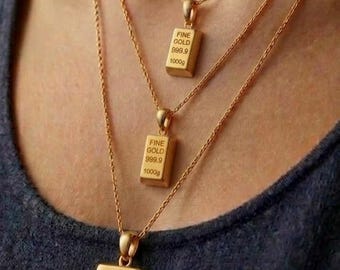 May include: Three gold-colored necklaces featuring rectangular pendants shaped like bullion bars. Each pendant is engraved with the text FINE GOLD 999.9 1000g. The necklaces are layered, with the smallest pendant at the top and the largest at the bottom.