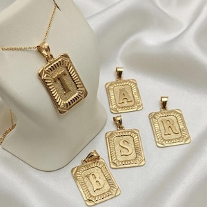 May include: A gold-toned necklace featuring a rectangular pendant with the letter T, accompanied by four additional rectangular pendants displaying the letters A, S, R, and B. Each pendant has a textured center and a decorative border.