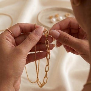 K&ouml;nnte beinhalten: Eine goldfarbene Halskette mit einer Kette und einem rechteckigen Gliederdesign. Die Halskette wird gerade geschlossen. Der Hintergrund ist ein weicher, cremefarbener Stoff. Weiterer Schmuck ist im Hintergrund zu sehen.