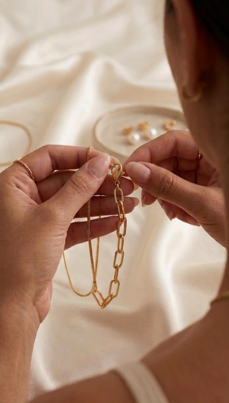 K&ouml;nnte beinhalten: Eine goldfarbene Halskette mit einer Kette und einem rechteckigen Gliederdesign. Die Halskette wird gerade geschlossen. Der Hintergrund ist ein weicher, cremefarbener Stoff. Weiterer Schmuck ist im Hintergrund zu sehen.