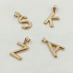 Pode incluir: Quatro pingentes de metal em tom dourado com o formato das letras S, K, Z e A. Cada pingente apresenta uma superfície lisa e arredondada com uma pequena argola integrada no topo para fixação, apresentados como componentes de joalheria.