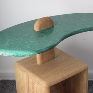 May include: A green and brown side table with a unique, organic shape. The top of the table is made of a green material with a speckled pattern. The base of the table is made of wood and has a natural finish.