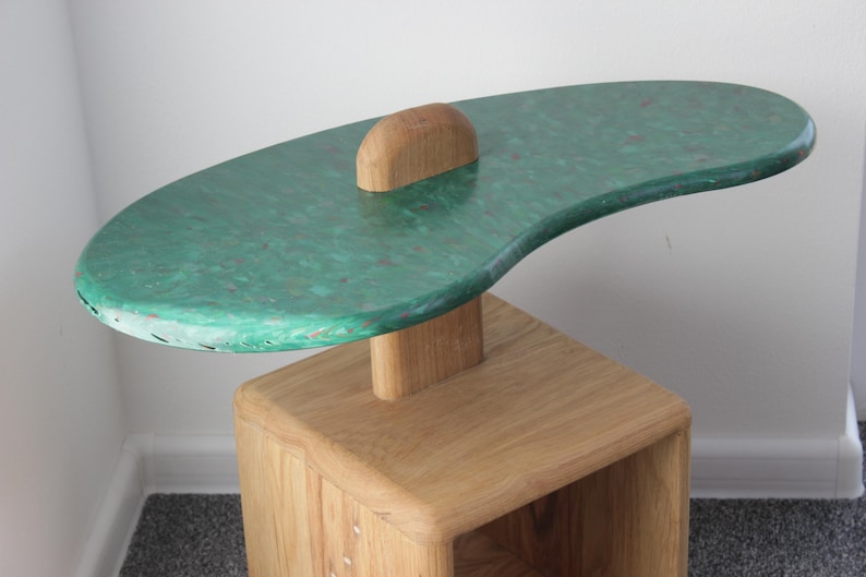 May include: A green and brown side table with a unique, organic shape. The top of the table is made of a green material with a speckled pattern. The base of the table is made of wood and has a natural finish.