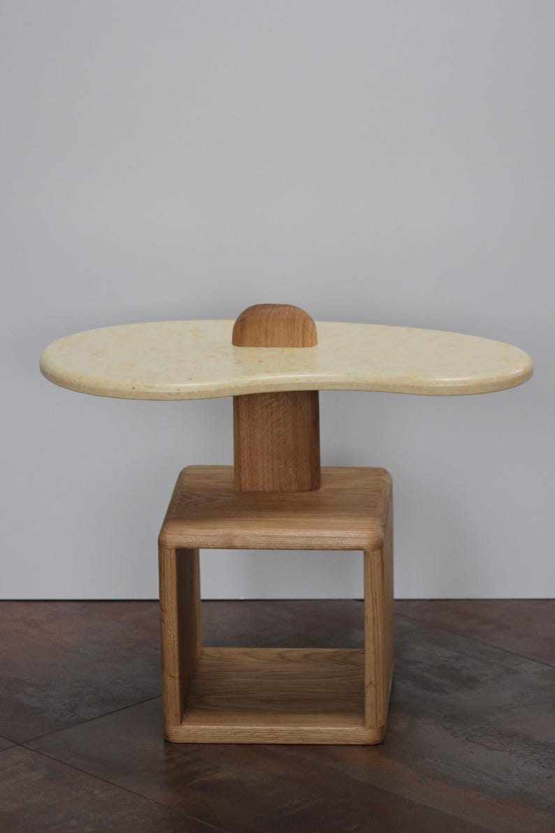 May include: A wooden side table with a unique, organic-shaped top and a square base. The top is a light cream color and the base is a natural wood tone.