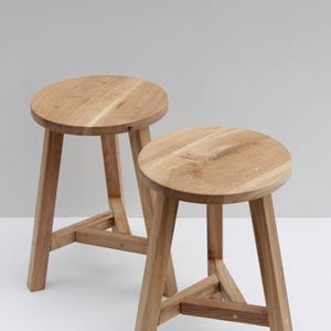 May include: Two light-coloured wooden stools with round seats and three angled legs. Each stool features a triangular support brace connecting the legs near the base. The wood grain is visible on the circular tops and the sturdy, straight-cut legs.
