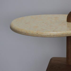 May include: Close-up of a light beige oval table top with a wood base. The table top has a speckled texture.