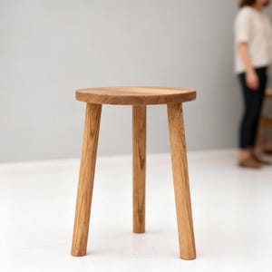 Può includere: Uno sgabello in legno a tre gambe con seduta rotonda e finitura naturale con venature del legno marrone chiaro. Lo sgabello presenta tre gambe cilindriche che si estendono verso l'esterno dal centro, offrendo una base stabile al mobile.
