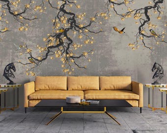 Puede incluir: Un mural de pared con ramas de árbol negras con flores doradas y pájaros pequeños sobre un fondo gris texturizado. Debajo, un sofá amarillo mostaza se sitúa tras una mesa de centro negra con patas doradas, flanqueada por cuatro mesas auxiliares con estatuas de caballos.